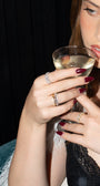 A woman with brown hair holds a glass of white wine, wearing a white feathered jacket over a black lace top, with a silver watch on her wrist against a dark background.